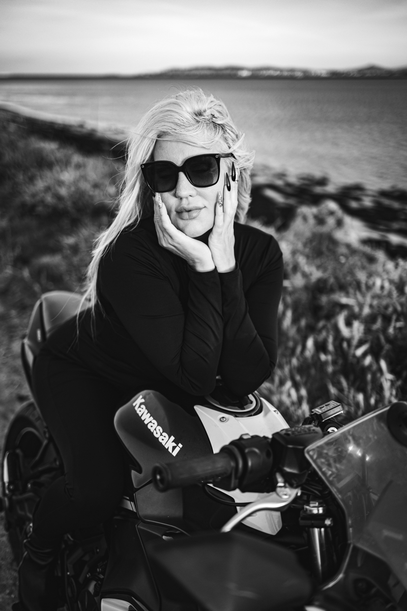 A woman with long blonde hair sitting on a Kawasaki motorcycle by the water, wearing black sunglasses and a fitted black outfit, resting her face on her hands.